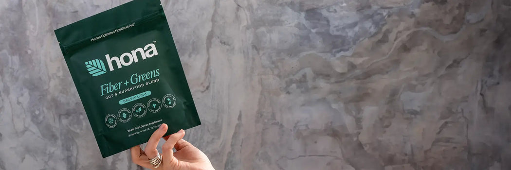 Hand holding a green package labeled Hona Fiber + Greens against a textured gray background.