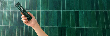 Hand holding a Hona product package against a green tiled wall
