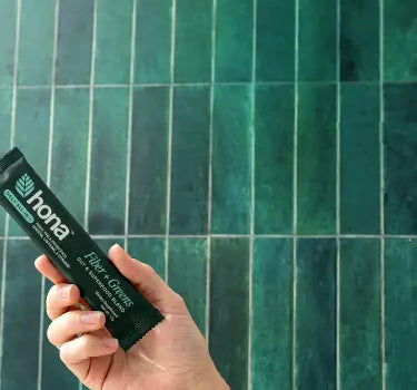 Hand holding a green tube of Hona gut health product against a green tiled wall.