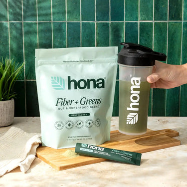 Hona Fiber + Greens package, shaker bottle, and stick pack on a wooden cutting board with a green tiled wall background.
